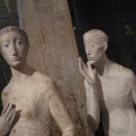Weiße Skulptur, bestehend aus zwei Menschen. Der Mann steht hinter der Frau ruft oder flüstert Ihr etwas zu.
