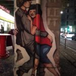Grafitti, welches eine junges Paar zeigt. Er ganz in grau mit Mütze hält sie (blaue, Hose, rotes Shirt, schwarzer Bob) im Arm.
