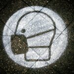 Eine auf den Bordstein gesprühter Hinweis, eine Maske zu tragen. Es handelt sich um einen weißen, gefüllten Kreis auf dem ein Mensch mit Maske abgebildet ist.